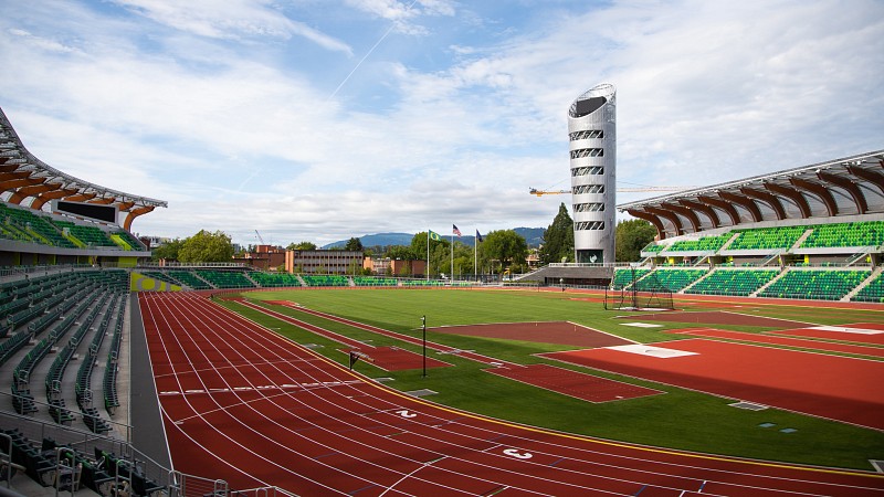 A wide shot of a track and field stadium on a sunny day. The green stadium seating and field are in the foreground. A large metal tower and bright blue sky in the background. 
